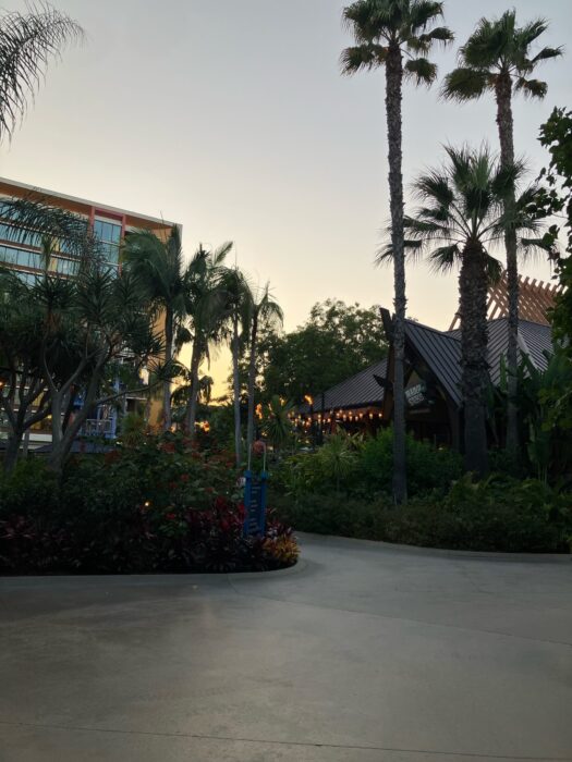 Photo of building housing Trader Sam’s Enchanted TIki Bar and Tangaroa Terrace Bar and Grill quick service dining location at Disneyland Hotel. Restaurants are surrounded by palm trees and tiki torches.