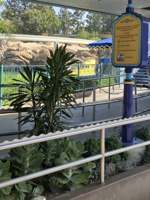 Photo showing queue area for Finding Nemo Submarine Voyage in Disneyland Park. Photo shows railings and chained delineating queue plus a sign with ride advisories among foliage. This photo illustrates the appearance of an outdoor queue at Disneyland Resort.