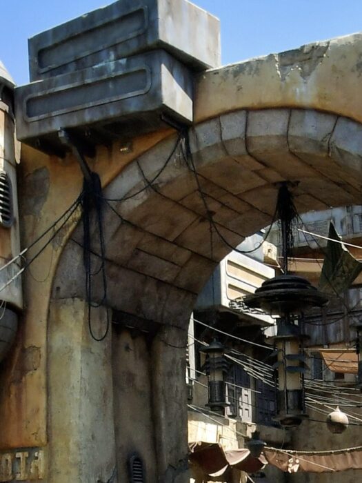 Photo showing archway in Star Wars: Galaxy’s Edge at Disneyland Park. Photo showcases the yellow and gray stone construction with lots of cargo boxes, hoses, pipes and hanging canopies and lighting.