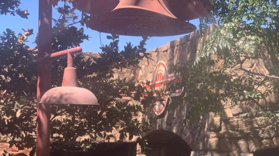 Photo of a fun line at Disneyland Resort showing a rock wall with wheel sign in queue for Radiator Springs Racers at Cars Land in Disney California Adventure Park, lamps and trees in foreground