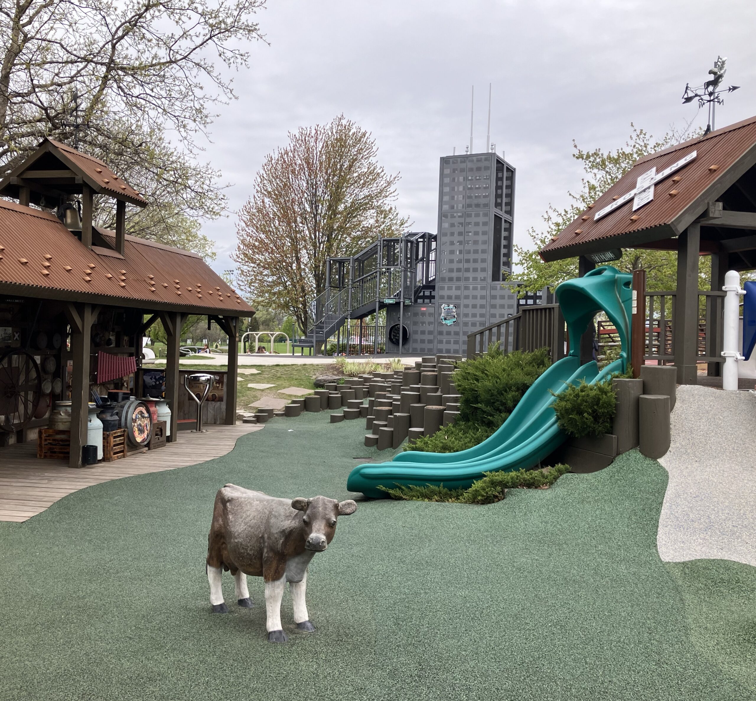 Photo of Harvester Park in Burr Ridge, Illinois. Shows a climbing structure resembling Sears / Willis Tower, a green slide, a open-sided barn structure with music equipment and a cow sculpture on a poured green play surface.