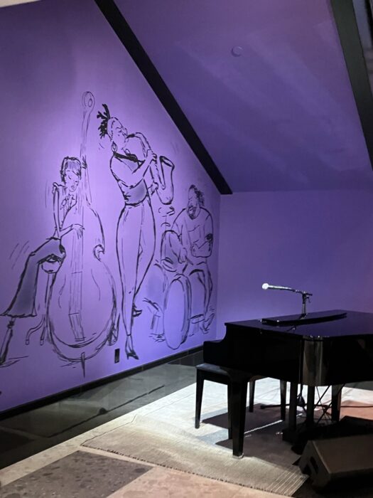 Photo showing lobby of Pixar Place Hotel where Joe Gardner character plays. Shows piano in a purple area under the stairs with paintings of musicians on the wall.