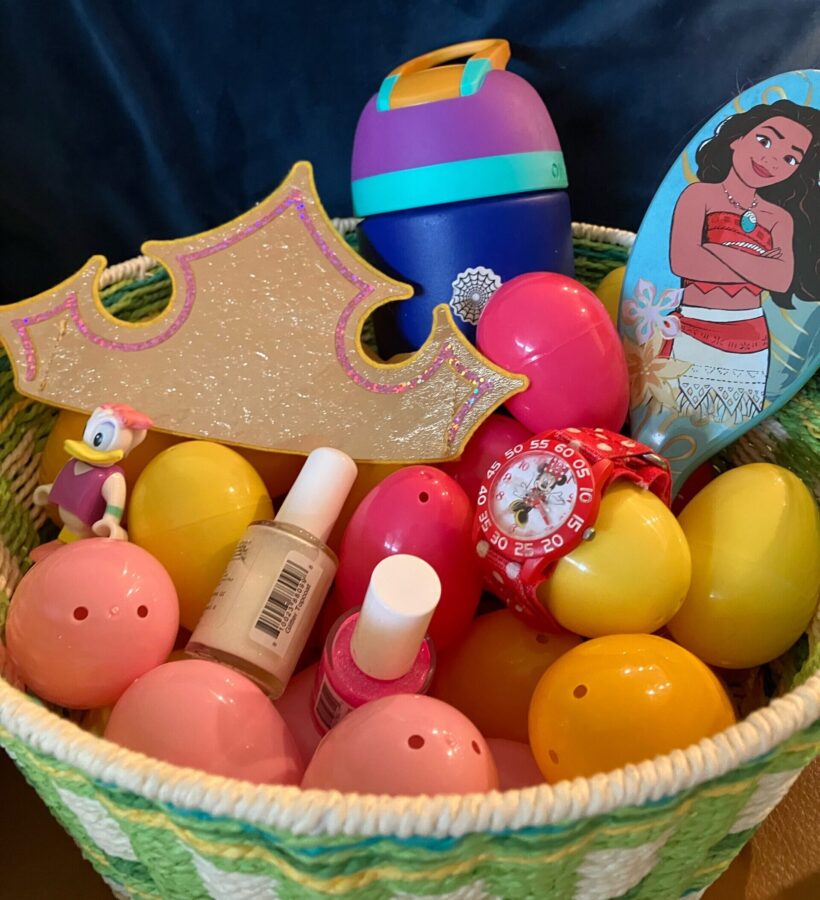 Photo of Easter basket with Disney themed egg fillers including a Moana hairbrush, a homemade princess crown, a Daisy Duck figure of a wooden railway, a Minnie Mouse watch, an Owala water bottle for a Disney trip and two bottles of glitter nail polish. The gifts are surrounded by Easter eggs in a basket.