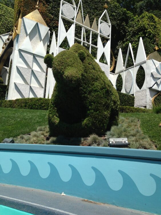 Photo of bear shaped topiary in front of “it’s a small world” facade at Disneyland Park in California. Attraction facade is geometric white and gold shapes in front of green hedges.