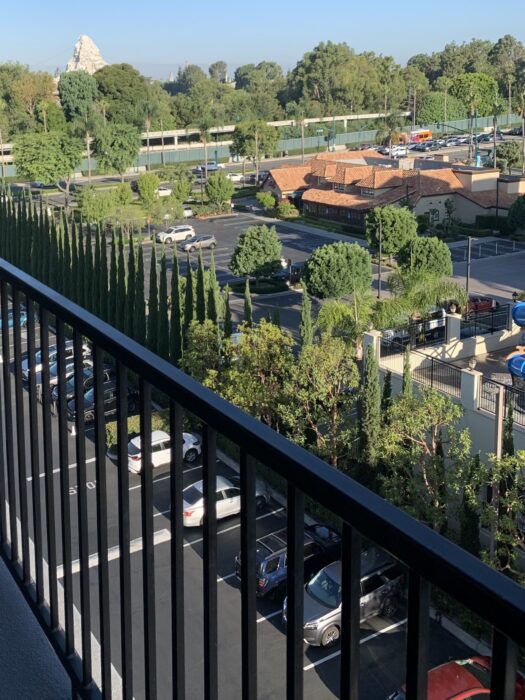 Photo showing the view from an outdoor walkway at the Fairfield Inn near Disneyland. There is a black iron railing, and over the railing is a lot of parking, Harbor Boulevard and Disneyland in the distance. At the top of the photo is Matterhorn Mountain at Disneyland.