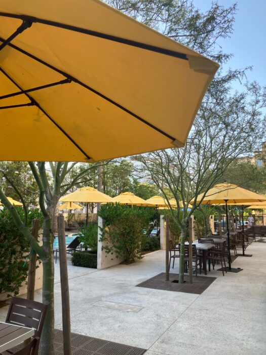 Photo showing seating around Home2 Suites Anaheim Pool Area. There are yellow umbrellas and small trees interspersed among tables and chairs.