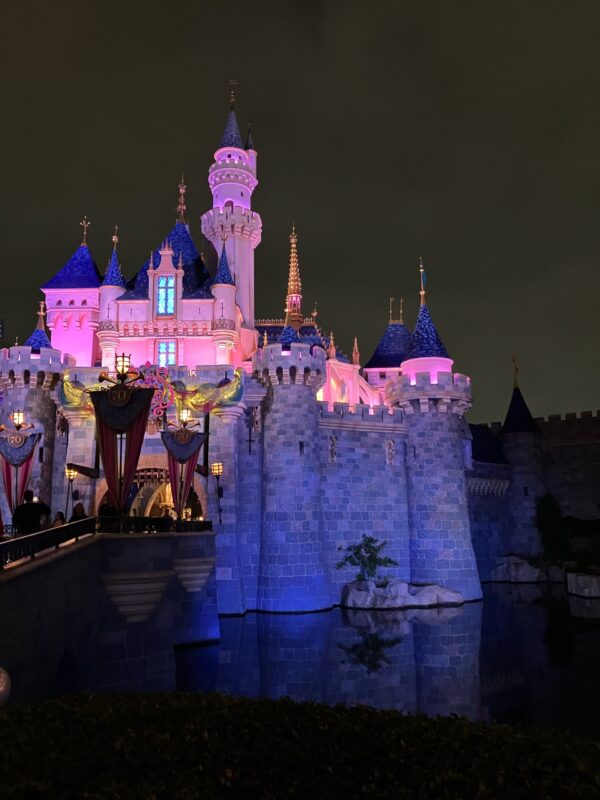 Photo of Disneyland’s Sleeping Beauty Castle taken at night. The castle is seen rising above the water and is lit with pink lighting.