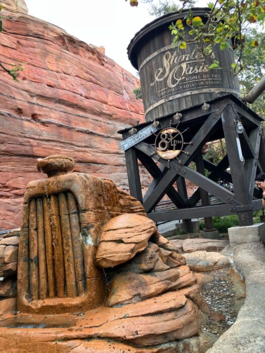 Photo showing prop from queue area for Radiator Springs Racers. Shows a wooden water tower in front a rocks shaped like an early automobile. In the background are read rocks and green foliage.