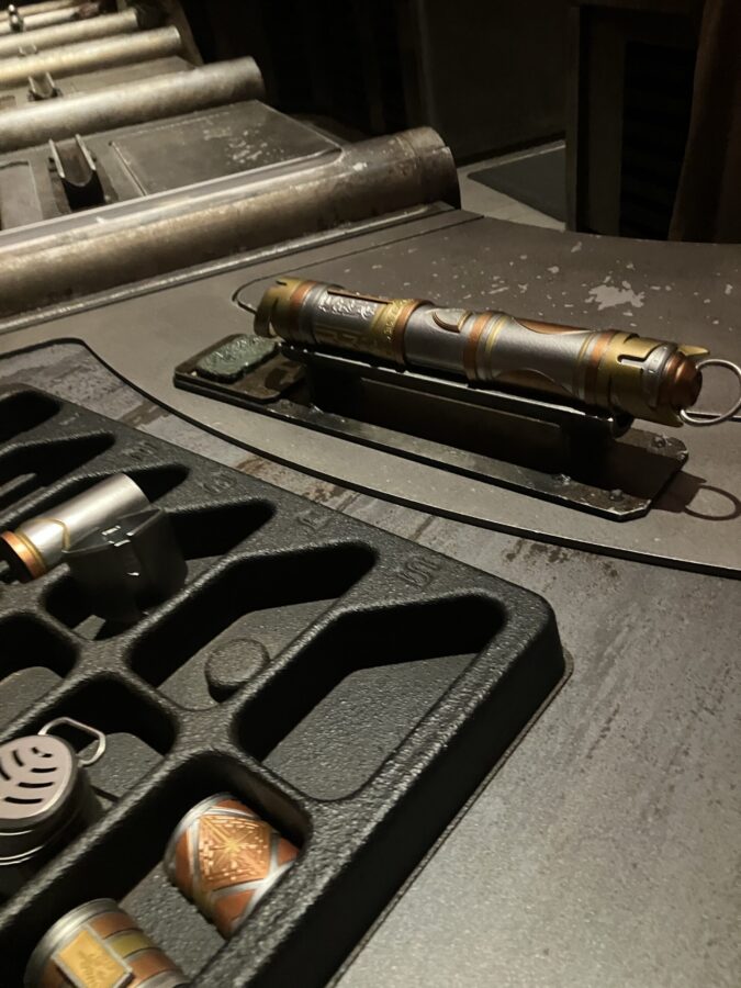 Photo taken inside Savi’s Workshop in Disneyland showing tray with open areas plus lightsaber parts. Behind the tray is an assembled lightsaber hilt.