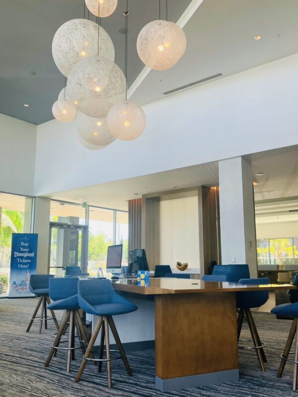 Image of the lobby at Marriott Element hotel in Anaheim, California near Disneyland Resort. Photo shows long high table with counter-height stools. Overhead is a large spherical light fixture and there is also a sign advertising Disneyland park tickets.
