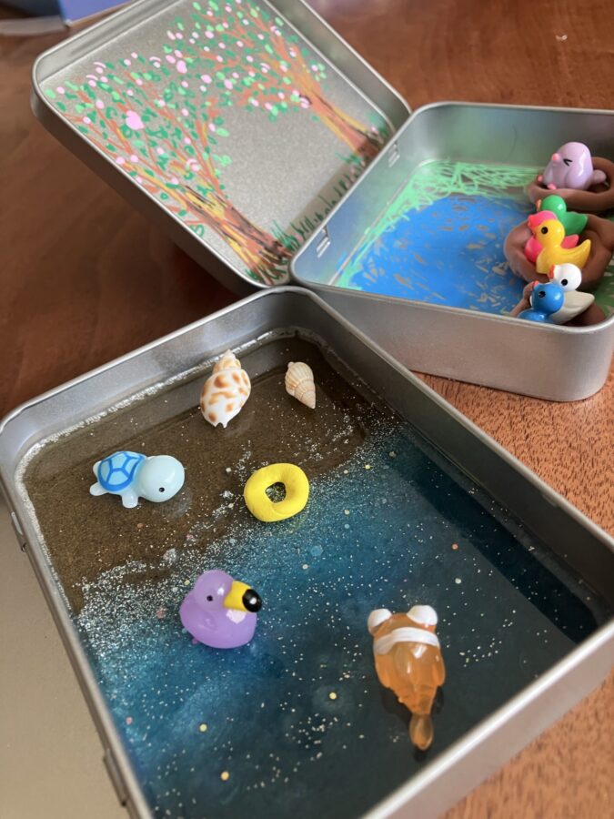 Photo of hinged tin boxes with little homemade play scenes.. One with small resin birds in brown clay nests. The interior of the tin is decorated with painted trees, pond and meadow. Beach playset with little resin animals, a clay inner tube and a back made out of resin, sand and blue mica powder.